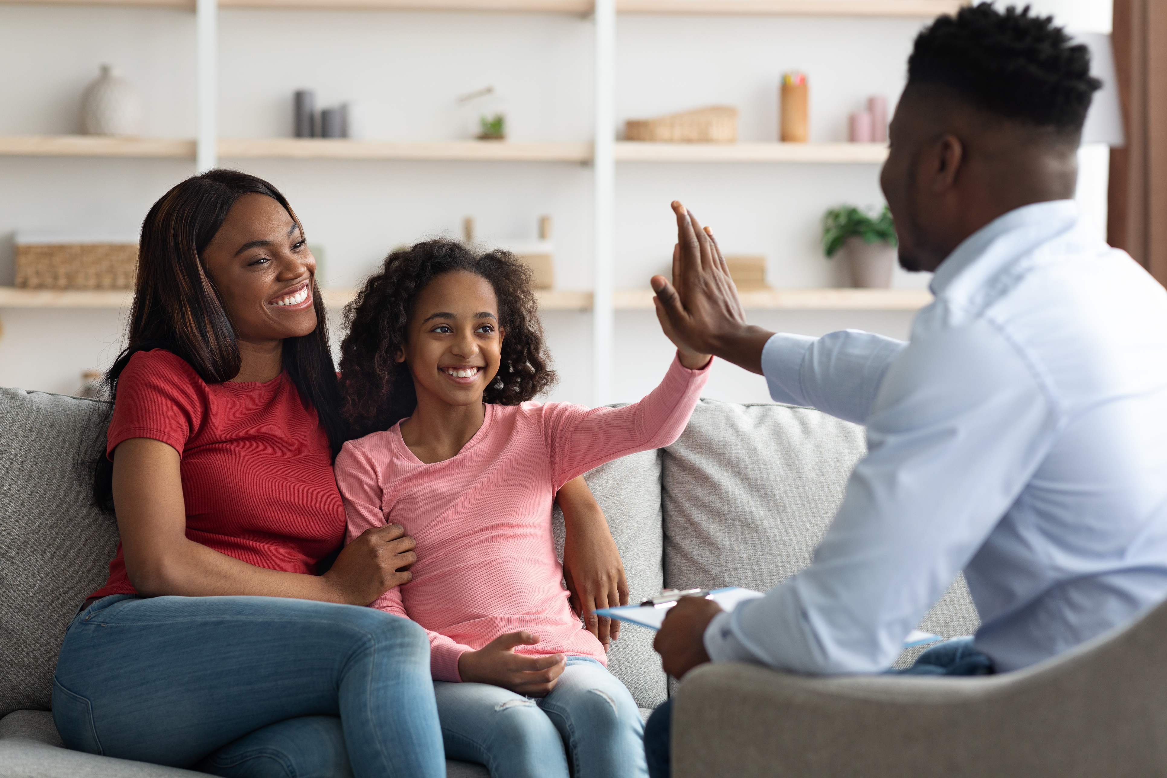 A family therapist high fiving a young girl sitting on a couch next to her smiling mother.